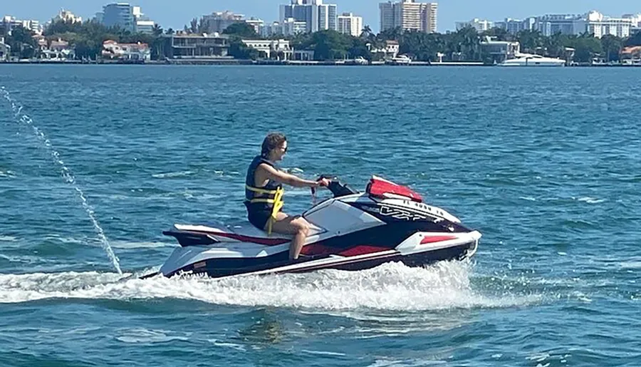 A person is riding a jet ski on a sunny day with a coastal city skyline in the background.