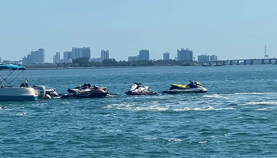 A group of personal watercrafts are floating near a boat against a backdrop of a coastal city skyline and calm blue waters.