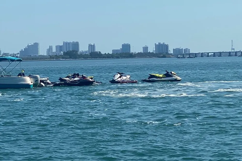 The image shows a collection of personal watercraft floating on the water with a boat and a city skyline in the background on a sunny day