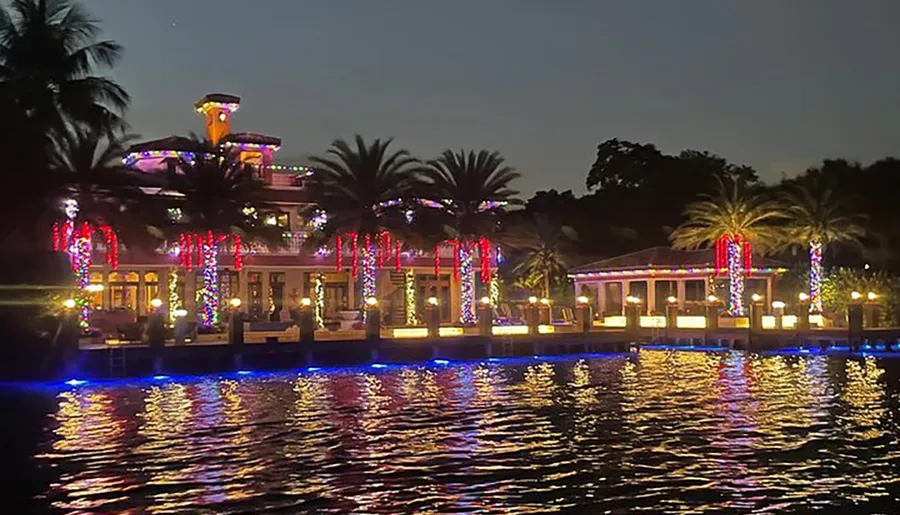 The image displays a waterfront building adorned with multicolored lights reflecting on the water's surface at twilight.