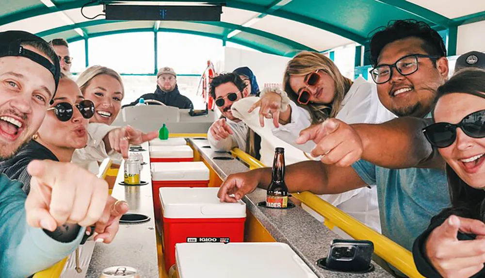 A group of cheerful people are pointing towards the camera while sitting at a bar with individual coolers with a bearded man in a cap driving the vehicle in the background
