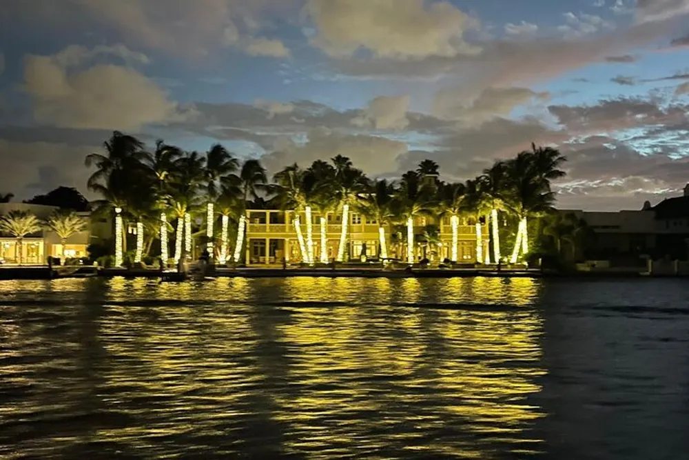 An opulent waterfront house is illuminated at night with lights reflecting off the water and silhouetting palm trees against the twilight sky