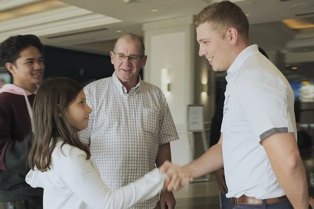 A young girl is shaking hands with a smiling young man while an older man and another young person are looking on in what appears to be a friendly and casual meeting