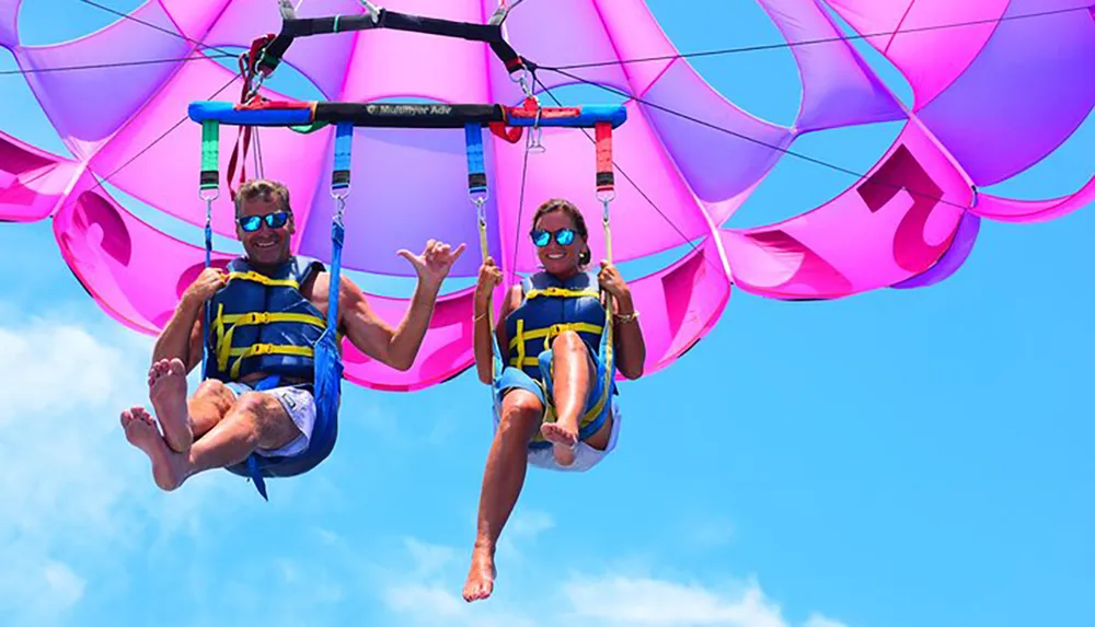 Two people are enjoying a parasailing adventure in a clear blue sky with a vibrant pink parachute above them