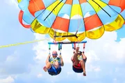 Two people are enjoying a parasailing adventure against a scenic backdrop of blue skies and fluffy white clouds.