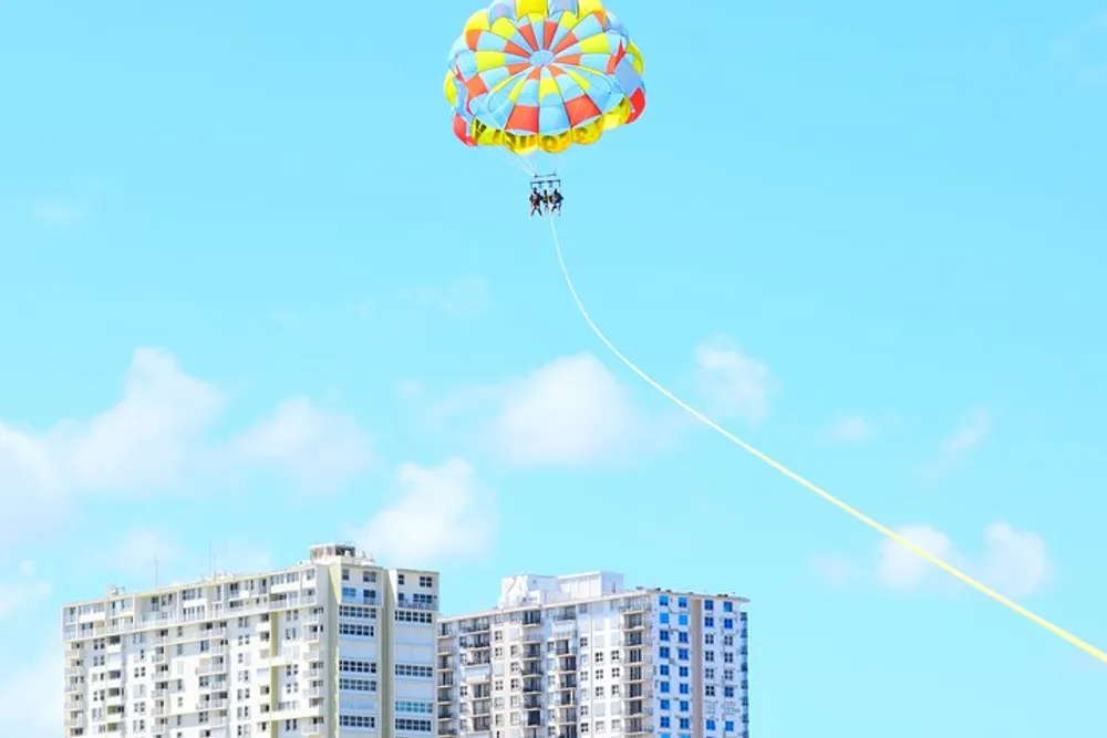 A colorful parasail with two people is flying high in the sky above apartment buildings on a sunny day