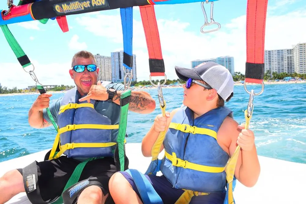 Two people are smiling while wearing life jackets and sitting on a parasailing boat ready for an adventure on a sunny day with a clear blue sky and coastal buildings in the background