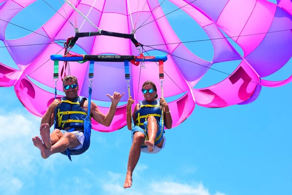 Two people are enjoying a parasailing adventure under a bright pink parachute against a clear blue sky