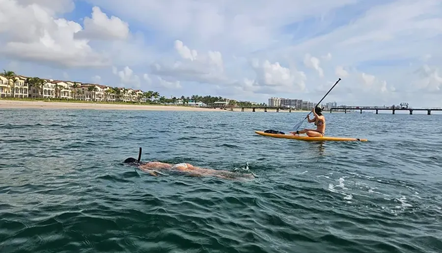 A person is paddleboarding near the shoreline while another person appears to be swimming or snorkeling in the sea nearby.