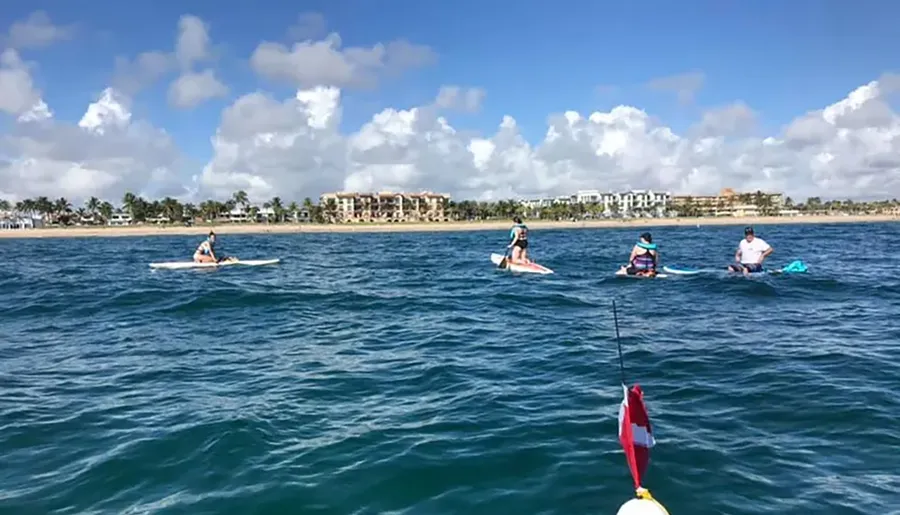 A group of people are paddleboarding on a calm blue sea near a coastal area with buildings in the background under a sky dotted with clouds.