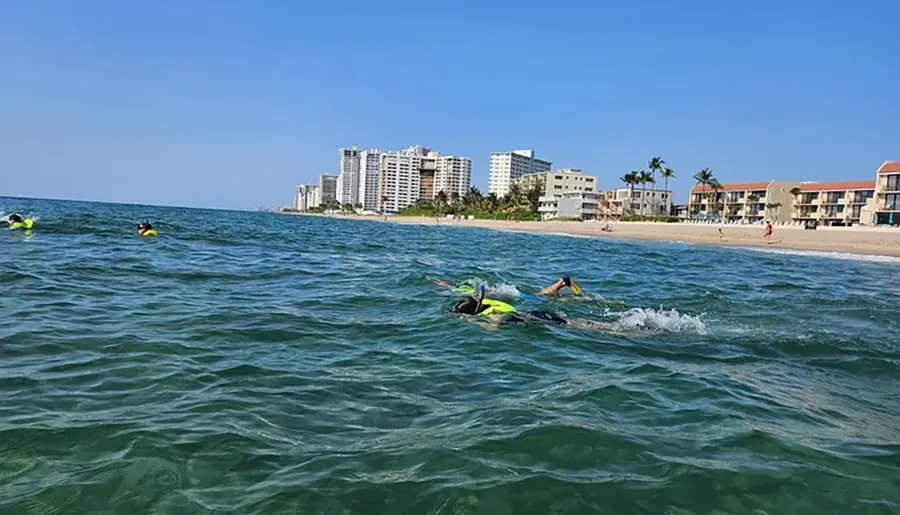 Swimmers wearing bright swim caps are seen in the ocean near a sandy beach lined with buildings on a sunny day.