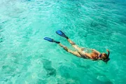 A person is snorkeling in clear turquoise water, wearing blue fins and an orange snorkel.