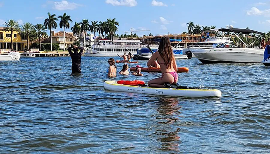A group of people are enjoying water activities on a sunny day, with some standing in the water, a girl sitting on a paddleboard, and luxurious houses in the background.