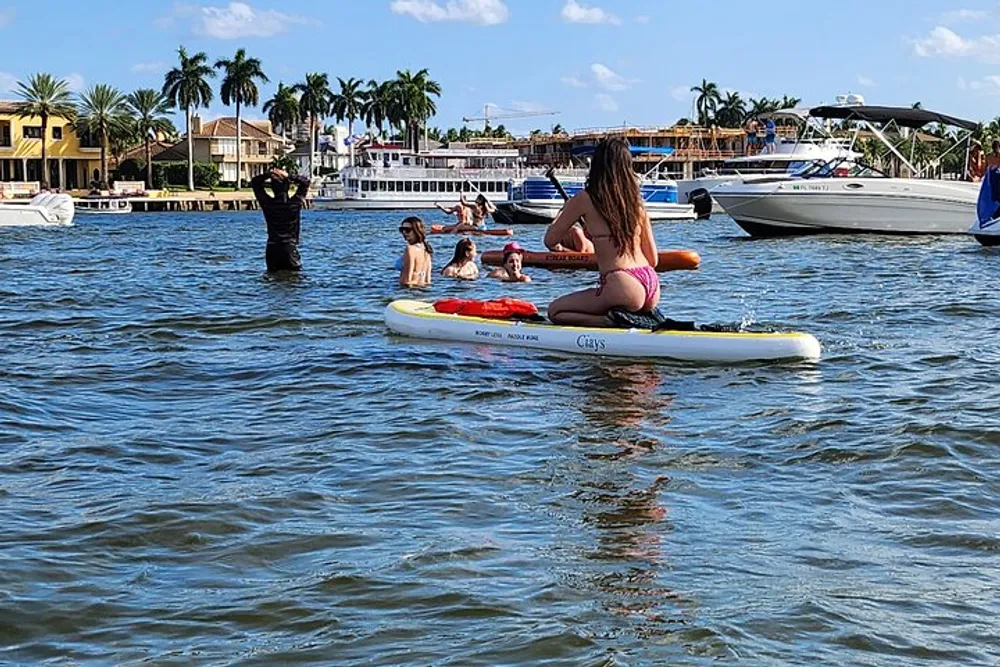 A person in a swimsuit sits on a paddleboard in a body of water with other individuals enjoying water activities in the background near waterfront houses