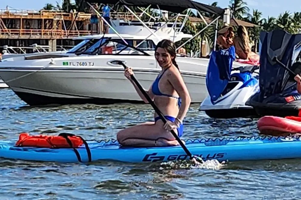 A person is kneeling on a paddleboard in a body of water paddle in hand with boats and jet skis nearby conveying a sense of aquatic leisure activities
