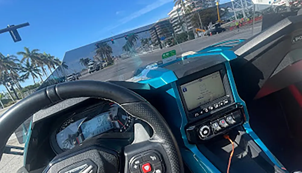The image shows the drivers view from inside a high-performance car with carbon fiber elements and digital screens stopped at a traffic light on a sunny day with palm trees in the background