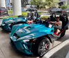 Two teal-colored three-wheeled vehicles are parked on a sunny day with four people wearing sunglasses sitting in them and several cars can be seen in the background