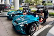 Two teal-colored three-wheeled vehicles are parked on a sunny day with four people wearing sunglasses sitting in them, and several cars can be seen in the background.