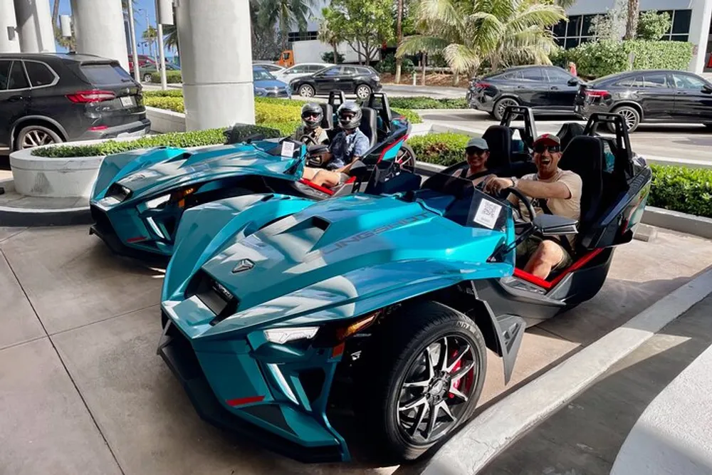 Two teal-colored three-wheeled vehicles are parked on a sunny day with four people wearing sunglasses sitting in them and several cars can be seen in the background