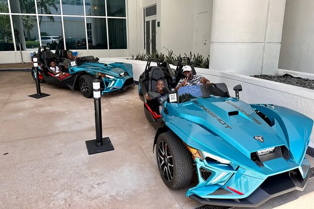 Two people are posing for a photo while sitting in a modern open-cockpit three-wheeled Polaris Slingshot vehicle which is parked outside a building