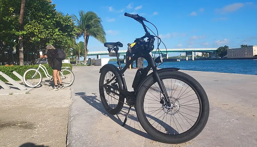 A black bike with thick tires is in the foreground on a sunny waterfront pathway, with a person standing near another bike and a bridge in the background.