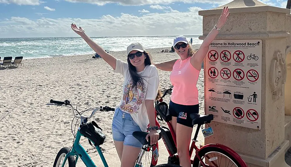 Two people are posing with raised arms next to their bicycles on Hollywood Beach near a sign listing beach regulations