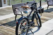 A black electric bike with a brown seat is parked on a wooden deck next to a metal bench, in daylight.