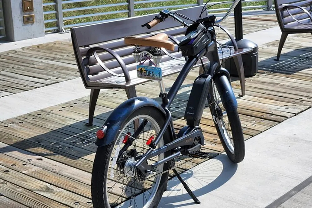 A black electric bike with a brown seat is parked on a wooden deck next to a metal bench in daylight