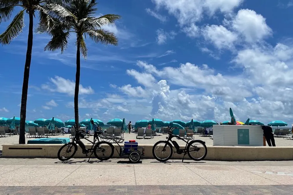 Two bicycles are parked on a sunny beach promenade lined with palm trees and vibrant turquoise beach umbrellas under a partly cloudy sky