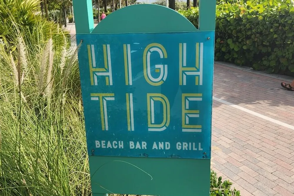 The image shows a turquoise sign with the words HIGH TIDE Beach Bar and Grill written in white and yellow letters suggesting it is a sign for a coastal dining establishment