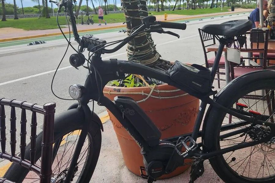 The image shows a black electric bike parked beside a large terracotta pot, with outdoor dining settings and a palm-tree-lined street in the background.