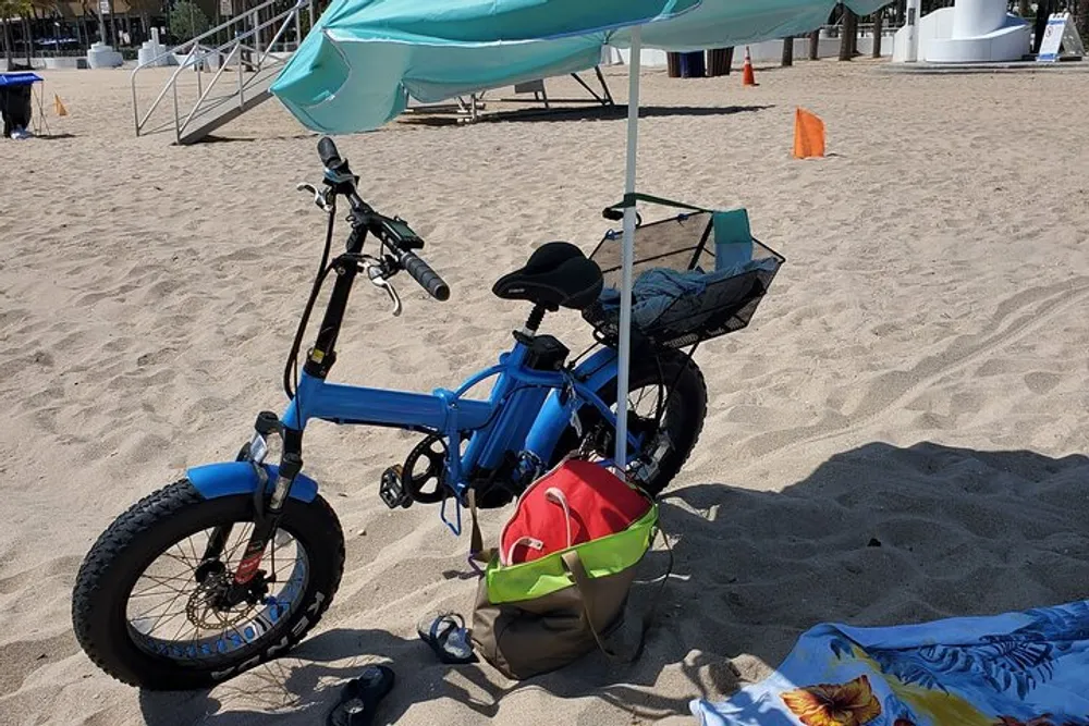 A blue foldable bike is parked on a sandy beach with a red bag hung on the handle a green tote bag beside it and beach accessories under an umbrella conveying a leisurely day by the sea