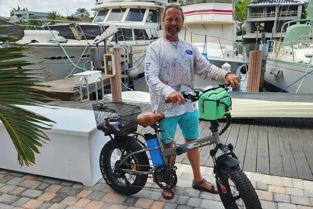 A smiling man stands next to a black electric bicycle with a basket and a green bag on the dock of a marina with boats in the background