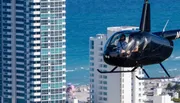 A helicopter with passengers is flying close to high-rise buildings near a beachfront.