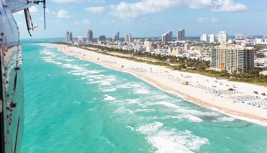 An aerial shot of a sun-drenched Miami beachfront lined with skyscrapers, umbrellas, and sunbathers, viewed from the side of a helicopter.