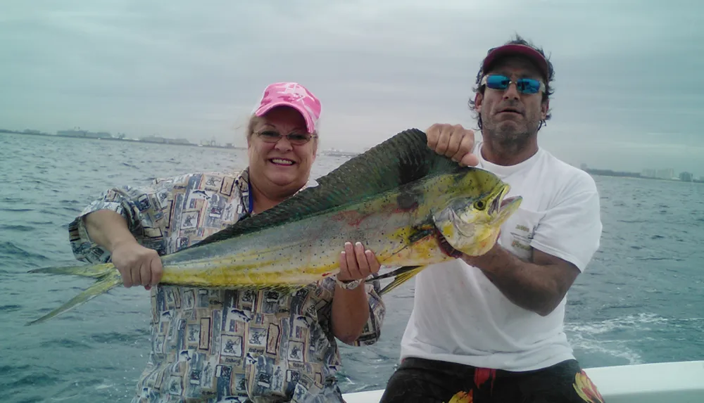 Two people are on a boat proudly displaying a large colorful fish they have caught with the ocean and a coastal city skyline in the background