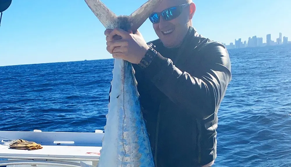 A man is smiling and holding up a large fish on a boat with the ocean in the background and a city skyline in the distance