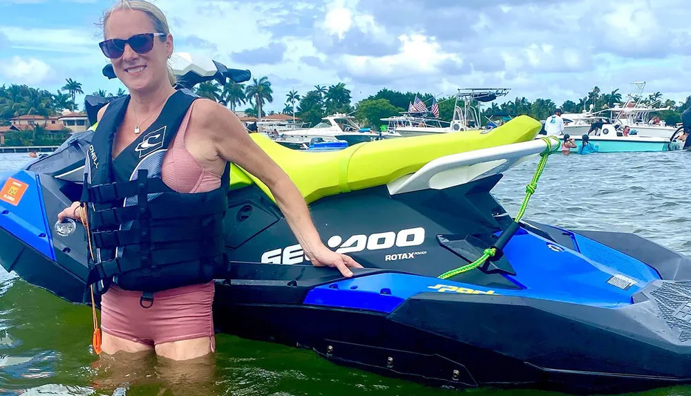 A person in a life jacket stands next to a jet ski on the water with other boats in the background