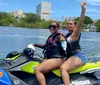 Two people are enjoying a jet ski ride on a sunny day with waterfront houses in the background