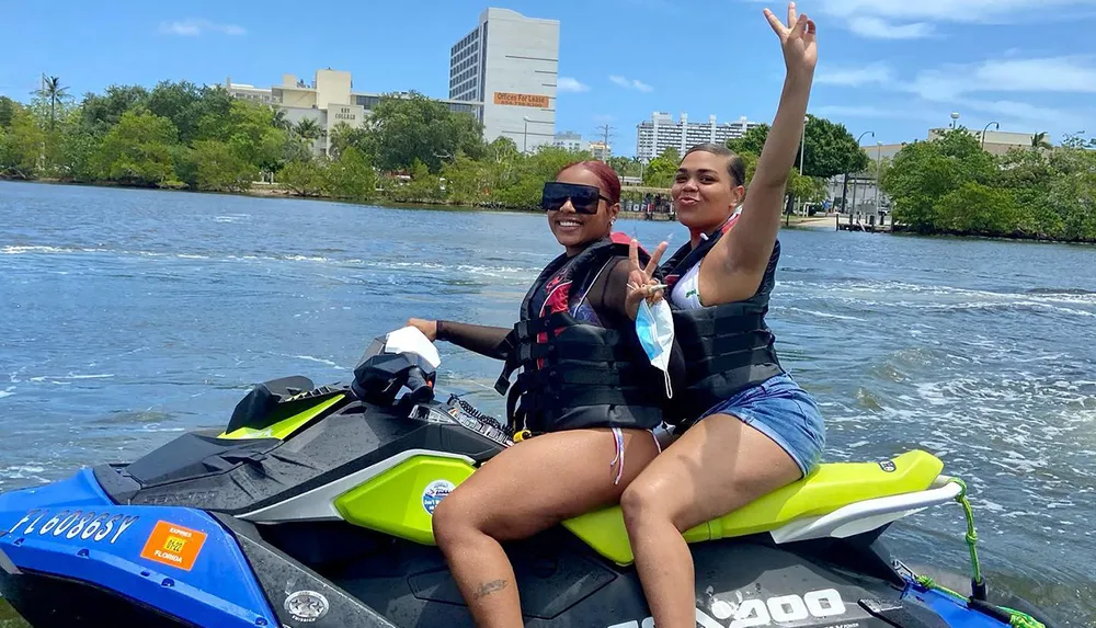 Two people are joyfully posing on a jet ski with one raising her arm in the air both wearing life jackets with a river and cityscape in the background