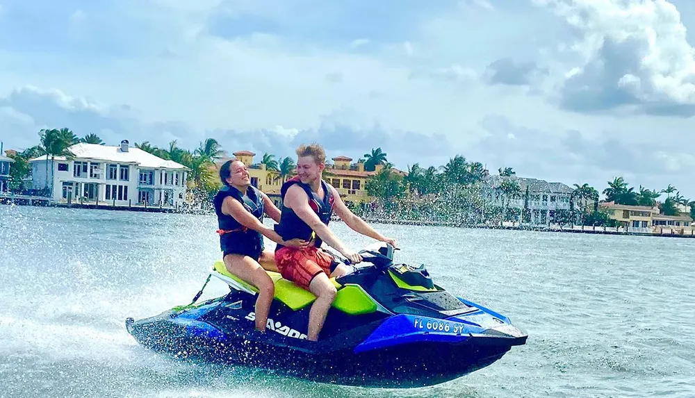 Two people are enjoying a jet ski ride on a sunny day with waterfront houses in the background