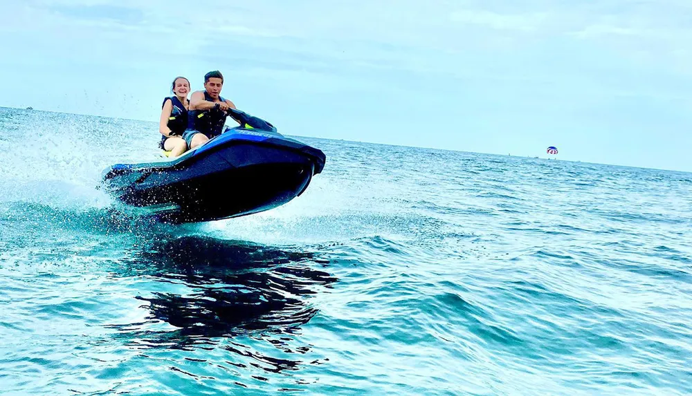 Two people are riding a jet ski jumping over a wave in the ocean with a clear sky above and a parasail in the distance