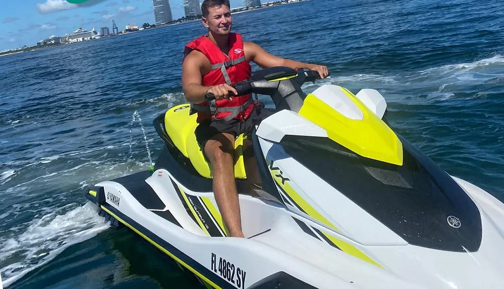 A man in a red life vest is riding a yellow and white personal watercraft on a sunny day with a coastal cityscape in the background