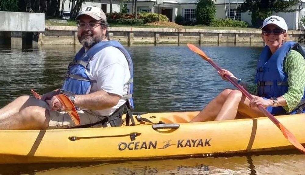 Two people are smiling while seated in a yellow tandem kayak on a sunny day