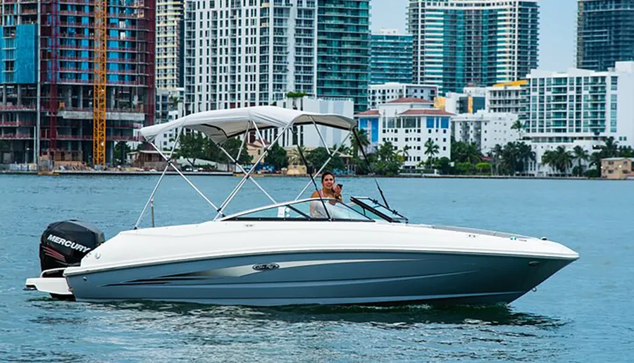 A person is steering a motorboat with a canopy on a body of water with a backdrop of modern, high-rise buildings.