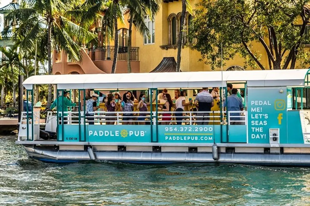 A group of people enjoys a social outing on a floating Paddle Pub boat cruising along a waterway with tropical scenery