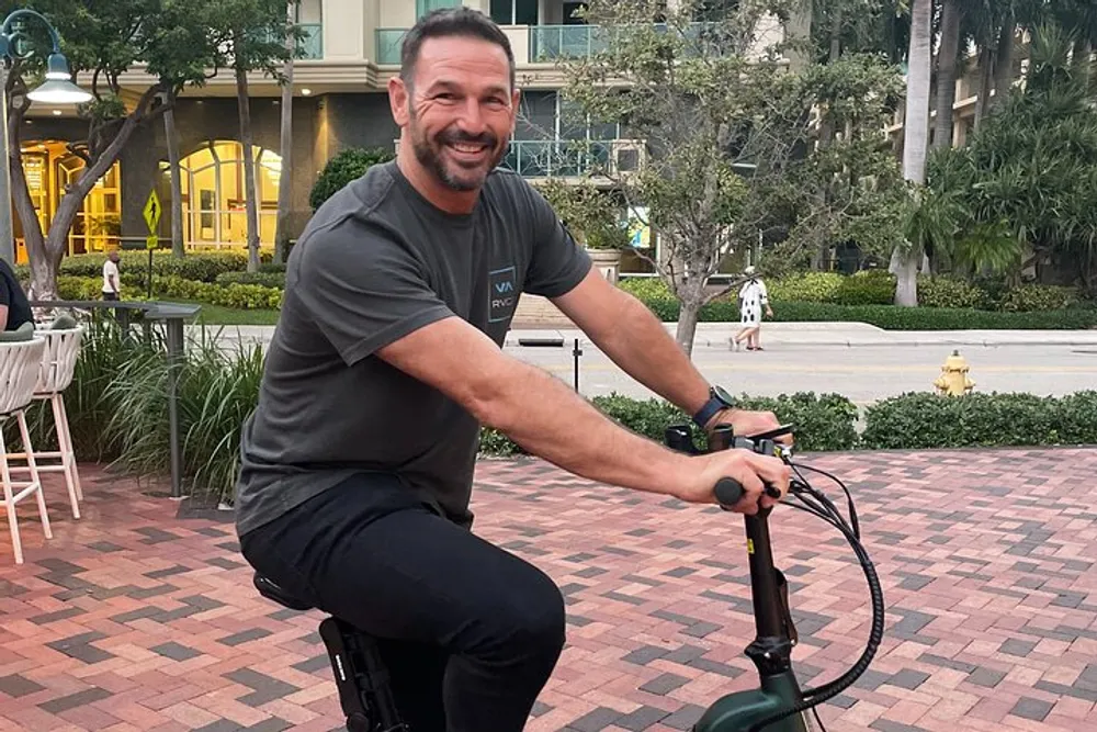 A smiling man is seated on a stationary bike outdoors with buildings and palm trees in the background