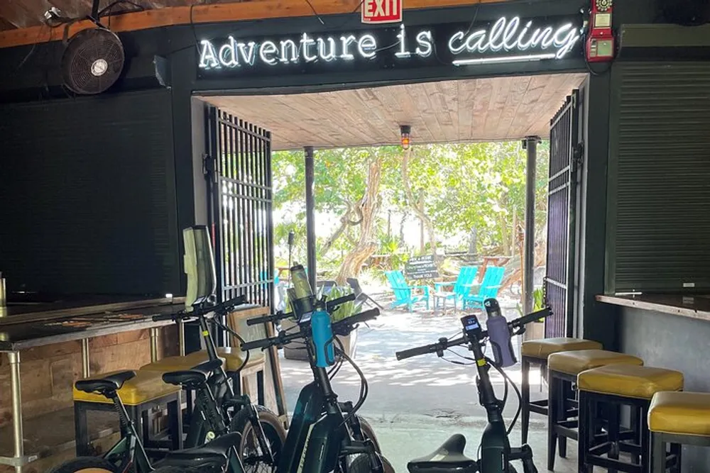 A neon sign reading Adventure is calling hangs above a row of stationary bikes inside a room that opens up to an outdoor area with colorful chairs