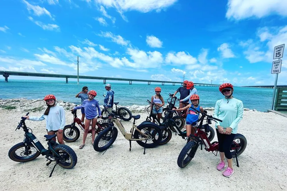 A group of people wearing helmets stands with their fat-tire bicycles on a coastal trail posing for a photo with a bridge and the ocean in the background