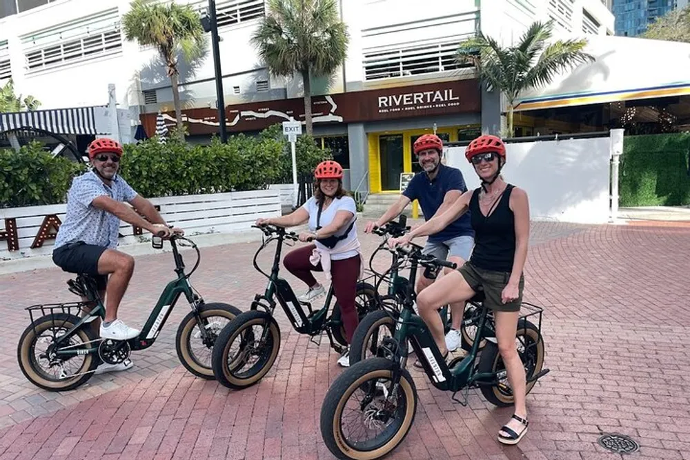 Four people wearing helmets are smiling for the camera while seated on fat-tire electric bikes in a sunny urban setting
